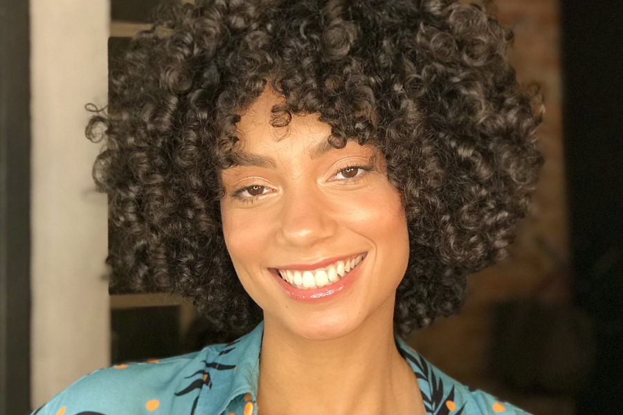 Isabela Reis é uma mulher jovem de pele negra clara, com cabelo black power curto, maquiagem simples e vestida de uma camisa azul