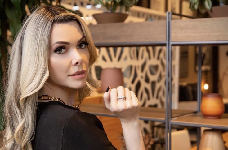 Doutora Miriane é uma mulher branca de cabelo com luzes loiras. Ela está vestida de preto, posando em diagonal para a câmera, com o braço flexionado para cima.