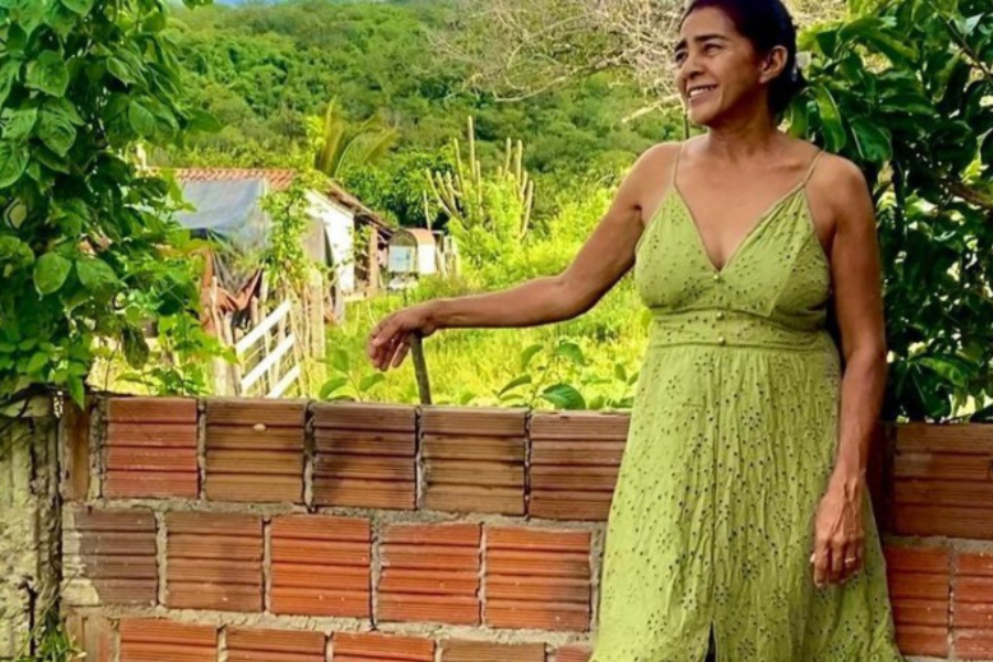 Maria Lúcia é uma mulher na faixa dos 50 anos de pele bronzeada. Ela sorri e usa um vestido longo verde. Está encostada em um muro de tijolos sem revestimento e, ao redor, há árvores e mato.